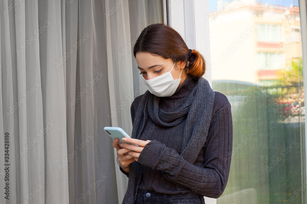 Woman in quarantine working at home, looking at her phone with a disposable medical mask on her face. Woman in quarantine working at home, looking at her phone with a disposable medical mask on her face.