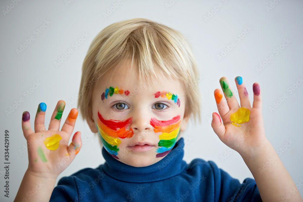 Beautiful blond toddler boy with rainbow painted on his face and messy hands Beautiful blond toddler boy with rainbow painted on his face and messy hands