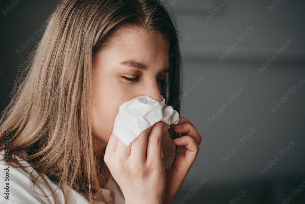 Young blonde white european blue-eyed woman at home on quarantine sneezes in a napkin on gray background. Coronavirus, illness, infection, quarantine, surgical bandage. Young blonde white european blue-eyed woman at home on quarantine sneezes in a napkin on gray background. Coronavirus, illness, infection, quarantine, surgical bandage.