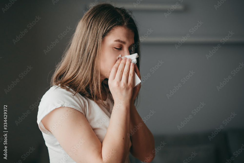 Young blonde white european blue-eyed woman at home on quarantine sneezes in a napkin on gray background. Coronavirus, illness, infection, quarantine, surgical bandage. Young blonde white european blue-eyed woman at home on quarantine sneezes in a napkin on gray background. Coronavirus, illness, infection, quarantine, surgical bandage.