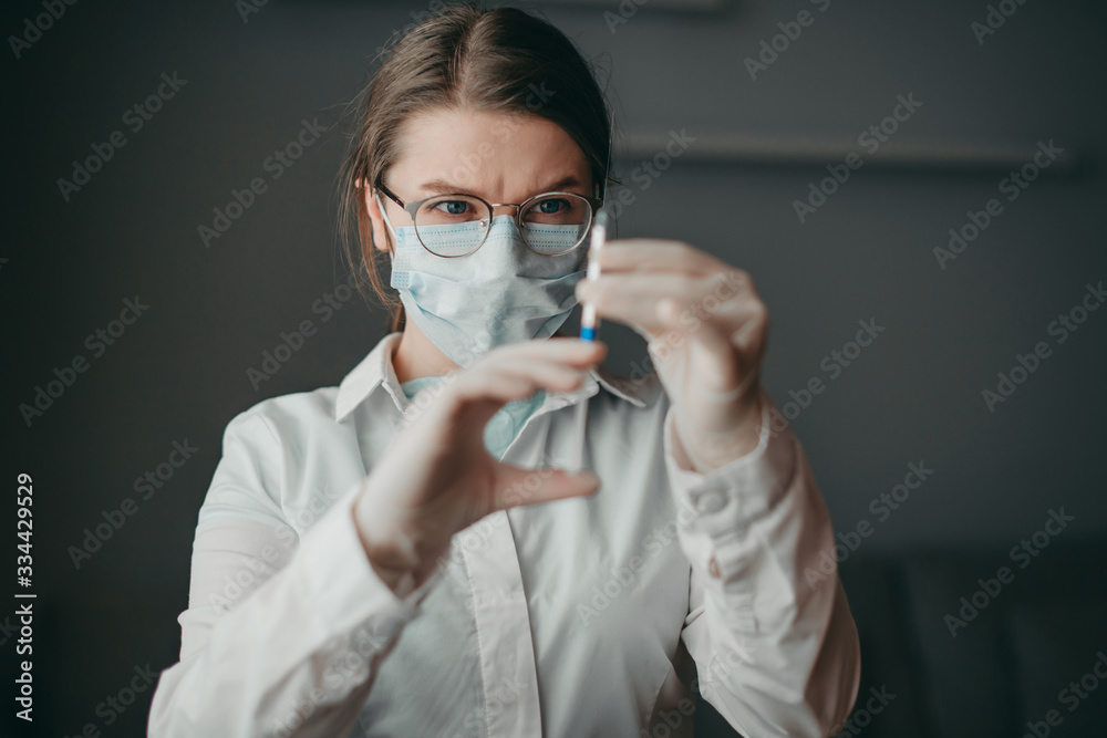Young blonde white european doctor woman with syringe in hand at hospital on quarantine with medical mask and glasses. Coronavirus, illness, infection, quarantine, flue, tablet, surgical bandage.