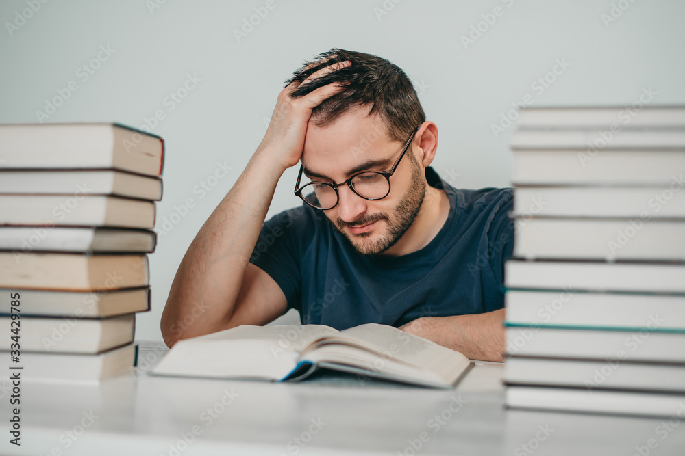 Young white European brunette man nerd in glasses teaches lessons, reads books, studied at university, prepares for exams, has many books at home in white backround table. Young white European brunette man nerd in glasses teaches lessons, reads books, studied at university, prepares for exams, has many books at home in white backround table.