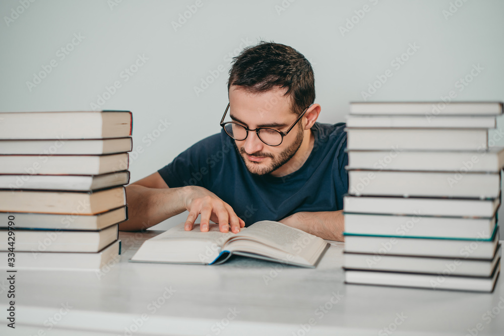 Young white European brunette man nerd in glasses teaches lessons, reads books, studied at university, prepares for exams, has many books at home in white backround table. Young white European brunette man nerd in glasses teaches lessons, reads books, studied at university, prepares for exams, has many books at home in white backround table.