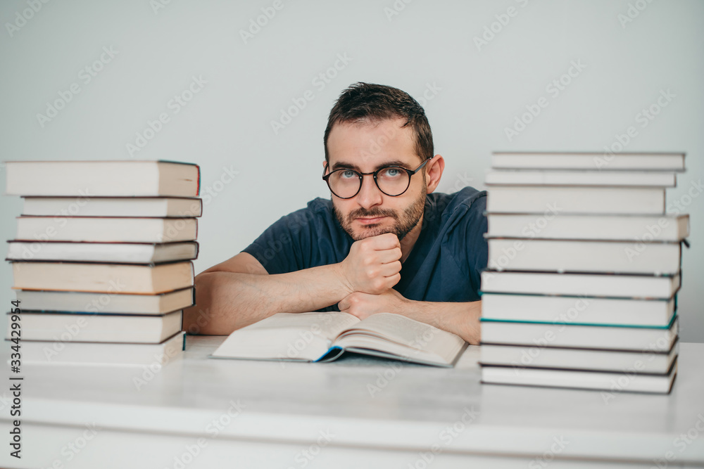 Young white European brunette man nerd in glasses teaches lessons, reads books, studied at university, prepares for exams, has many books at home in white backround table. Young white European brunette man nerd in glasses teaches lessons, reads books, studied at university, prepares for exams, has many books at home in white backround table.