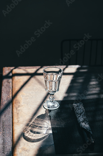 A glass goblet on a leg stands on a table among the shadows