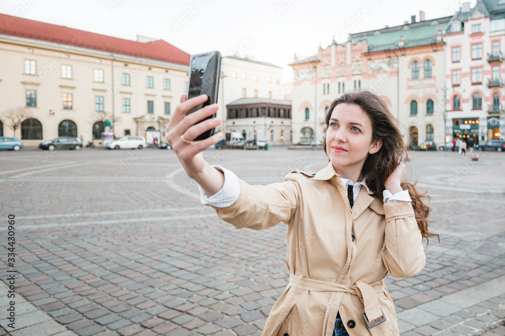 Fototapeta premium Happy young woman taking selfie at city street. Happy tourist girl walking outdoors. Spring fashion portrait of pretty brunette female posing in old town.