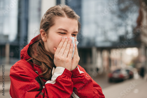 Young blond european woman sneezes in a napkin in the city on a background of modern architecture. Disease, protection, coronavirus, virus, disease, flu, respiratory dressing, allergy. 