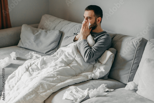 Young sick bearded european man sneezes into napkin at home on gray sofa with white blanket. Disease, protection, coronavirus, virus, disease, flu, respiratory dressing.