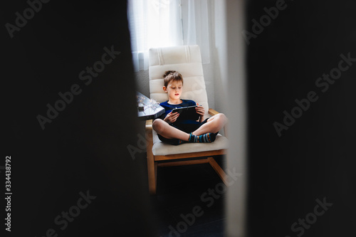 Young boy sitting in armchair and holding a tablet. Spying through the door opening. Daily technology for playing and learning. Consept of children using mobile devices.