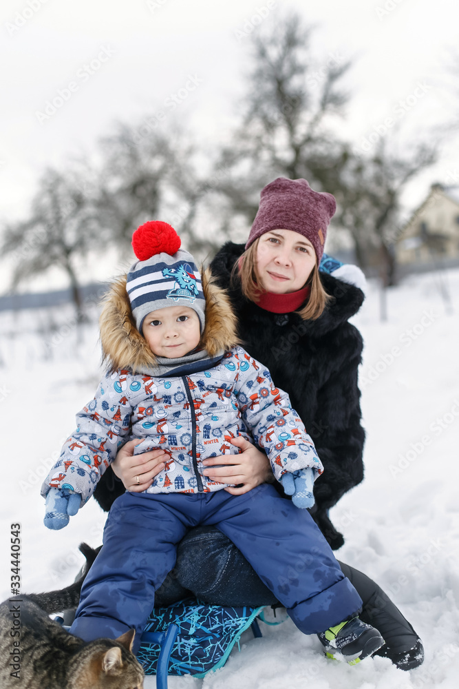 Smilling mother and her son sitting in the sledge with a striped cat in the winter garden.