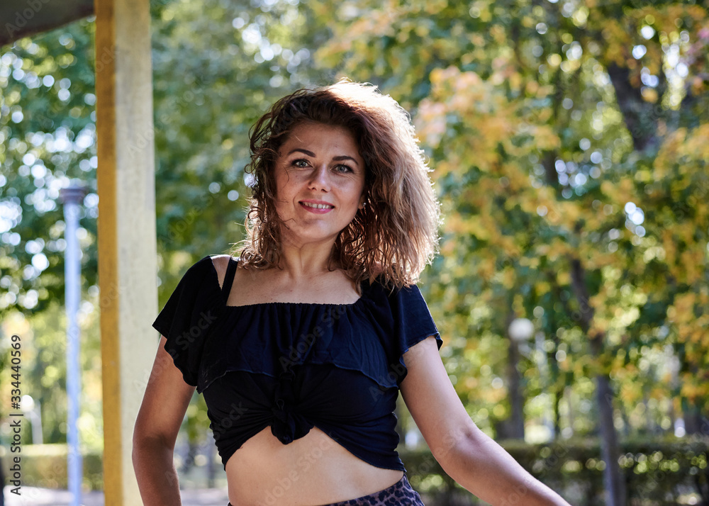 Young brunette woman, wearing black top, leather jacket and dark long skirt and high heels, posing near yellow rusty fence in city park. Urban lifestyle female photo session in summer autumn spring. Young brunette woman, wearing black top, leather jacket and dark long skirt and high heels, posing near yellow rusty fence in city park. Urban lifestyle female photo session in summer autumn spring.
