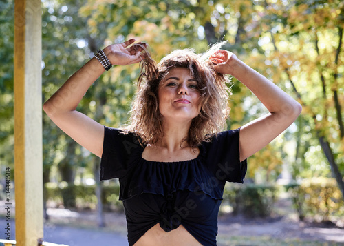 Young brunette woman, wearing black top, leather jacket and dark long skirt and high heels, posing, touching curly hair with hands in city park. Urban lifestyle female photo session in summer autumn