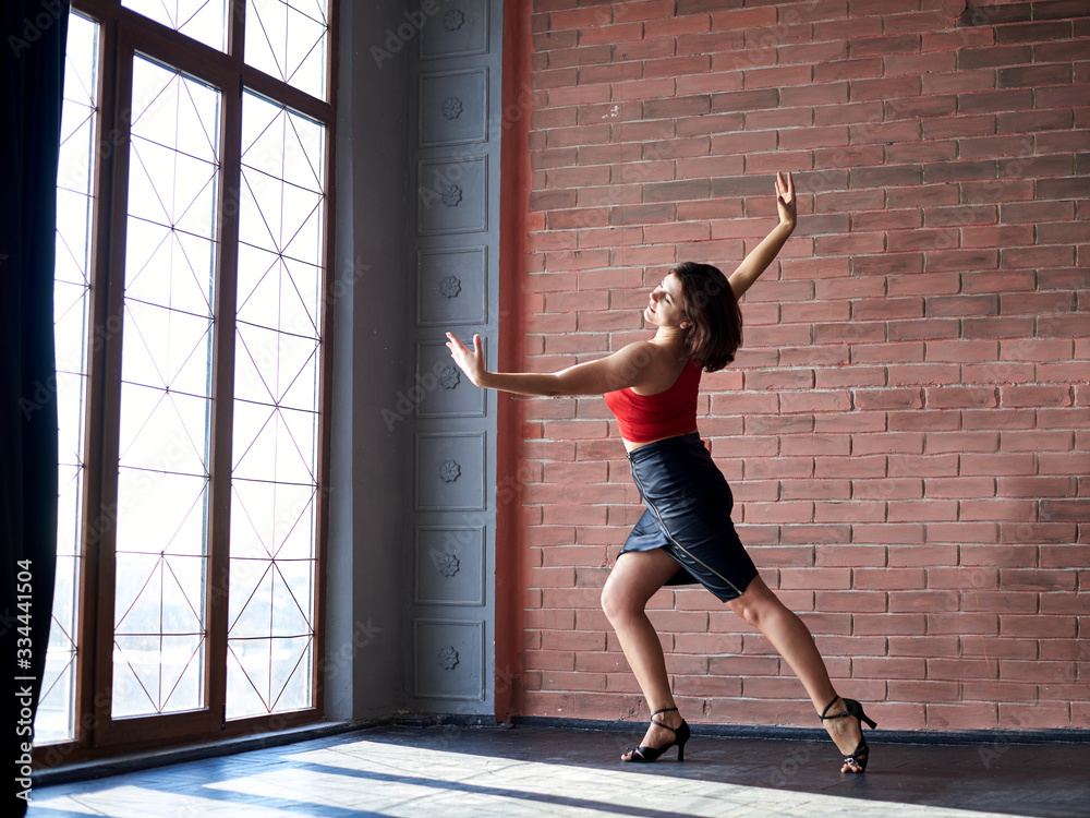 Young brunette woman, wearing red top and black leather skirt, dancing posing in spacious studio loft room with huge window. Bachata salsa dancer, training in light apartment with brick wall.