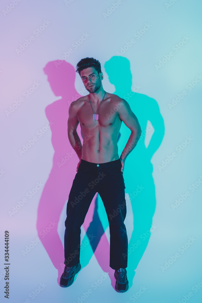 full length view of sexy, confident man in dark blue jeans holding hands on hips and looking at camera on blue background with shadows full length view of sexy, confident man in dark blue jeans holding hands on hips and looking at camera on blue background with shadows
