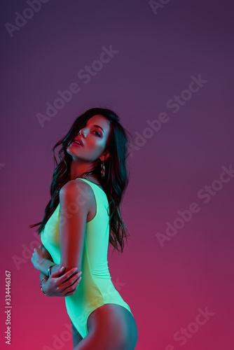 seductive girl in neon green swimsuit posing at camera with crossed arms on purple background with gradient