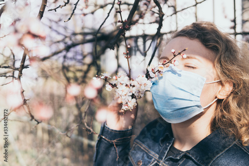 Young blonde woman smells cherry tree blossom through medical mask. Conceptual image of corona virus quarantine and allergy