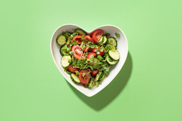 Fresh salad with tomato, cucumber, vegetables, microgreen radishes in plate shape of heart on green. View from above. Concept vegan and healthy eating.