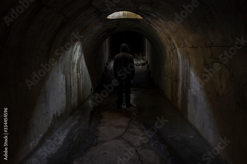 The silhouette of the figure of a man going into unknown darkness in a gloomy dark old underground passage lit by sunlight through a hatch in the ceiling.
