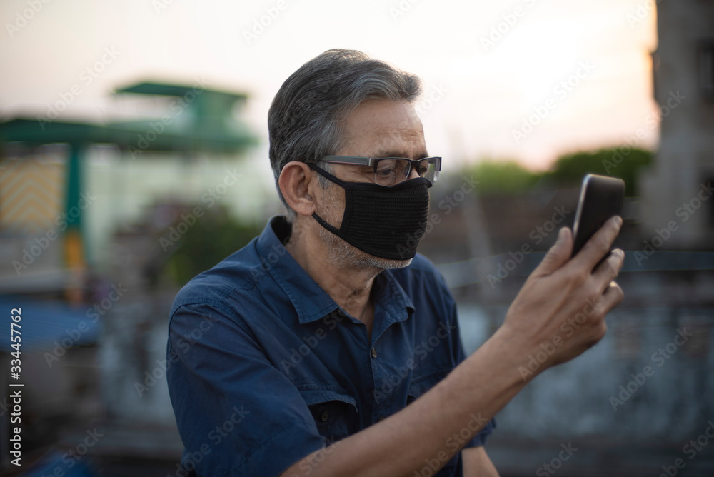 Portrait of an Indian old man wearing corona preventive mask in home isolation watching his mobile on rooftop. Indian lifestyle, disease and home quarantine. Portrait of an Indian old man wearing corona preventive mask in home isolation watching his mobile on rooftop. Indian lifestyle, disease and home quarantine.