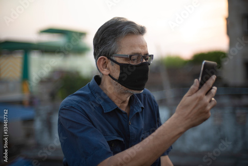 Portrait of an Indian old man wearing corona preventive mask in home isolation watching his mobile on rooftop. Indian lifestyle, disease and home quarantine.