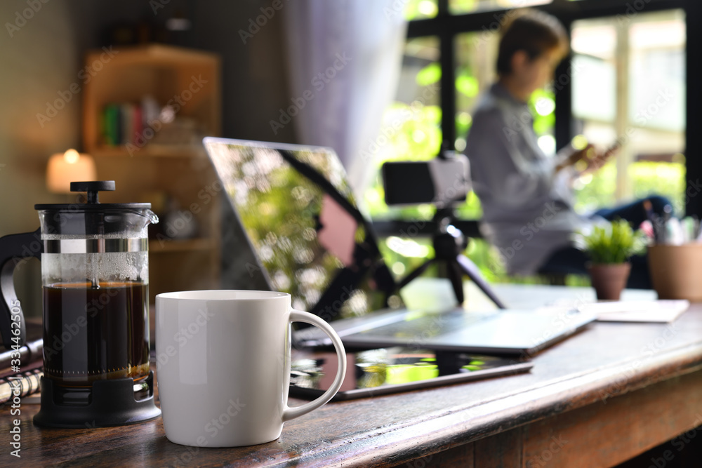 A coffee press and a cup on a work desk with a person working from home A coffee press and a cup on a work desk with a person working from home