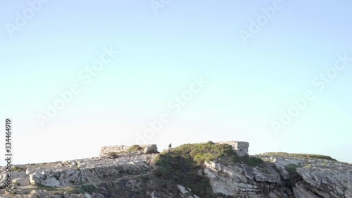 Distant man walking on a small peninsula with ruins, and the camera panning to the right unraveling the sea.