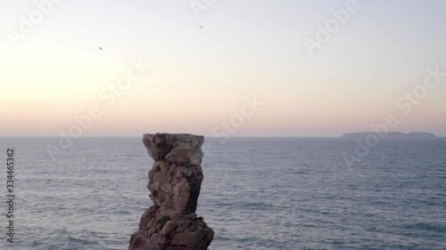 Large rock structure with seagulls flying over the top in the sunset with the horizon in the ocean in the background.