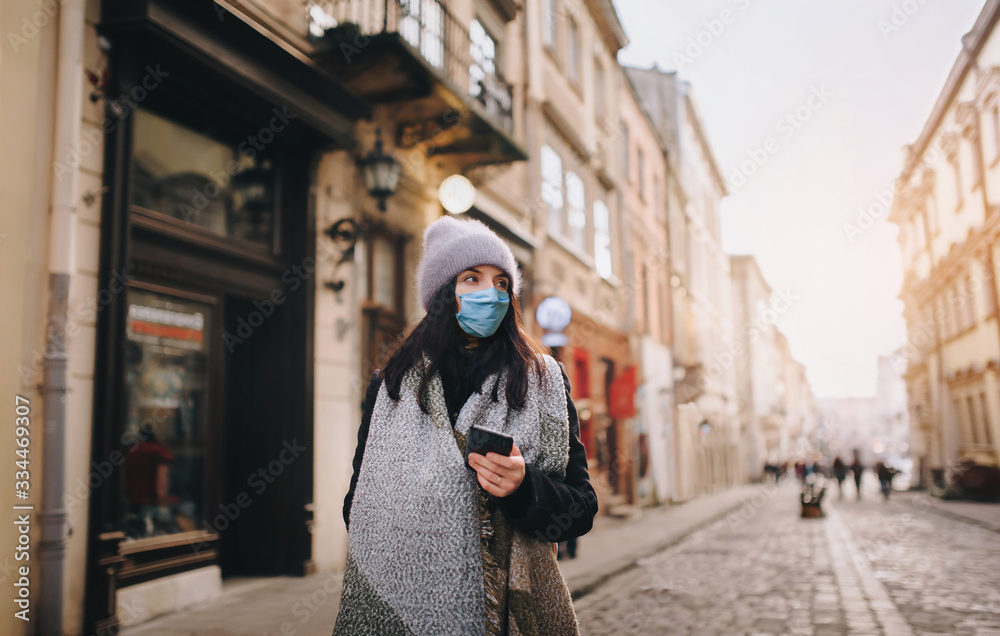 A young girl in a hat, a coat of a protective blue medical mask-respirator with a smartphone in her hand walks along the evening street of a deserted city. Quarantine, coronavirus, pandemic COVID-19. A young girl in a hat, a coat of a protective blue medical mask-respirator with a smartphone in her hand walks along the evening street of a deserted city. Quarantine, coronavirus, pandemic COVID-19.