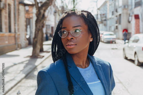 Close up portrait of a beautiful young african american woman with pigtail hairstyle in a blue jacket and glasses smiling and walking along the street © Nana_studio