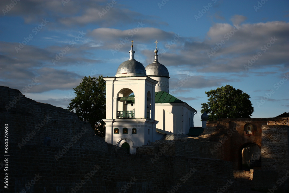 Ancient Russian city of Izborsk. Ancient stone fortress. Beams of the evening sun light white walls, a belltower and domes of St. Nicholas Church.