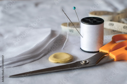 Needle and pin on white thread rolls for hand sewing on white cloth with tape measure and sketch yellow chalk with scissors cut on the fabric close-up.