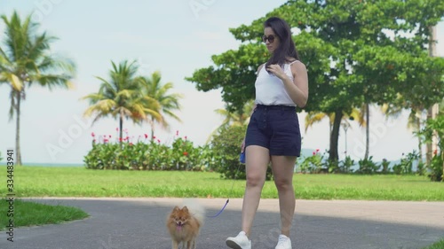 young woman walking her adorable small dog in city park. Beach, green lawn and trees