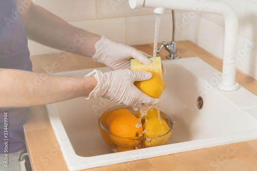 A man washes a lemon with a detergent in gloves. A young man sterilizes fruits and vegetables during an epidemic. Hands in disposable gloves close-up wash a lemon under running water.