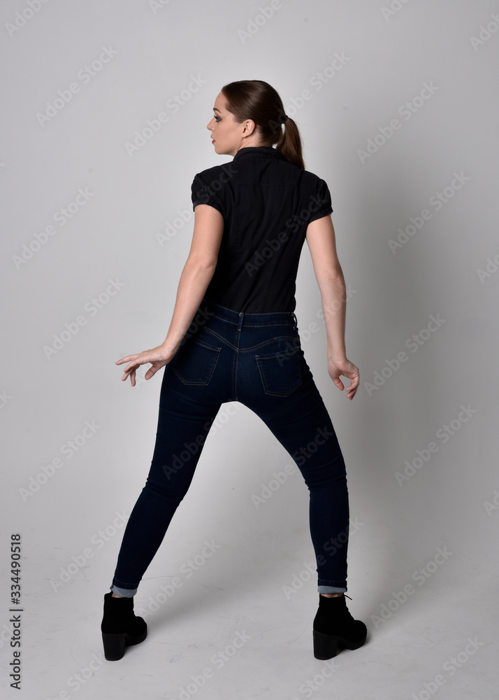  Portrait of a pretty brunette girl with ponytail, wearing jeans boots and a black blouse.  full length standing pose, with back to the camera on a studio background.