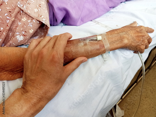 The hand of a man holding  wrinkled arm of an elderly woman with adhesive tape around her arm and syringe on her arm on a white bed cloth at the hospital.