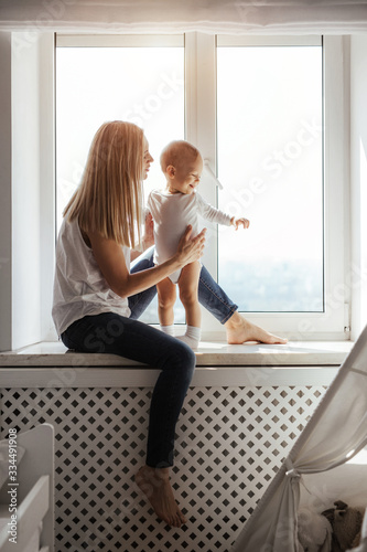 Portrait of a smiling young cute mother and her son playing near the window in a bright white room. Sunny day outside. True mother love