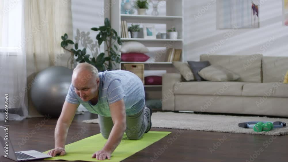 Senior man in sportswear doing upward facing dog exercise on mat in the living room while practicing yoga at home with video lesson on laptopSenior man in sportswear doing upward facing dog exercise o Senior man in sportswear doing upward facing dog exercise on mat in the living room while practicing yoga at home with video lesson on laptopSenior man in sportswear doing upward facing dog exercise o