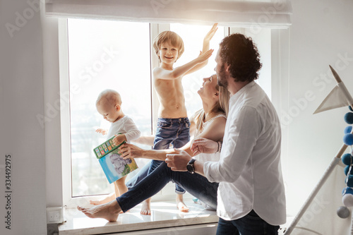 Happy smiling modern caucasian family playing and spending time in a bright white room near the window. Sunny day. Perfect parenting relationship. Parents of two cute boys.