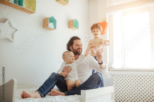 Young father of two sons playing with a toy plane. Happy family, three boys in a bright sunny room spending time together. Big brother hugs father