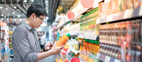 Asian man choosing orange juice in supermarket using smartphone to check shopping list. Male shopper with shopping cart selecting beverage bottle product in grocery store.