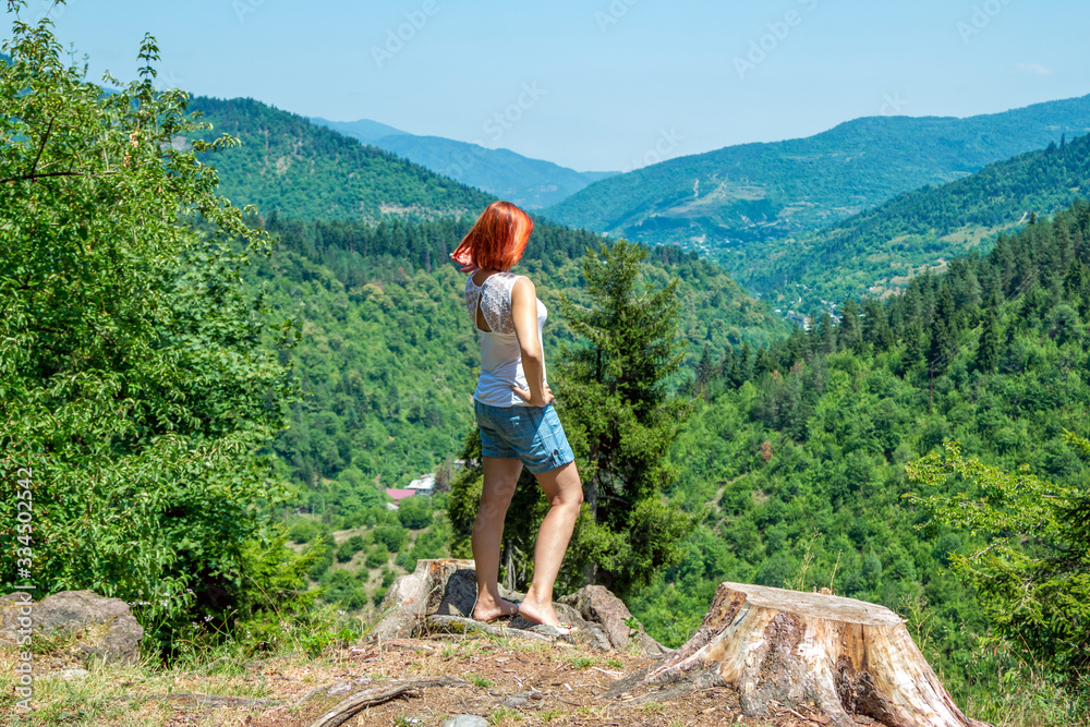 Beautiful woman with red hair, wearing blue jeans shorts, standing on the top of the hill, observing summer landscape - mountains, covered with green trees. 