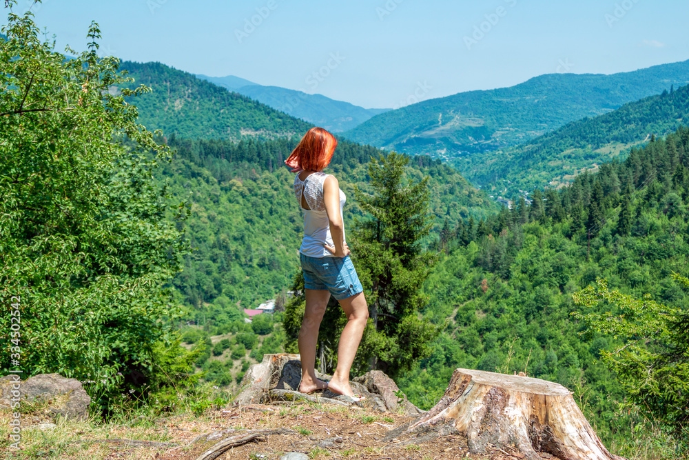 Naklejka premium Beautiful woman with red hair, wearing blue jeans shorts, standing on the top of the hill, observing summer landscape - mountains, covered with green trees. 