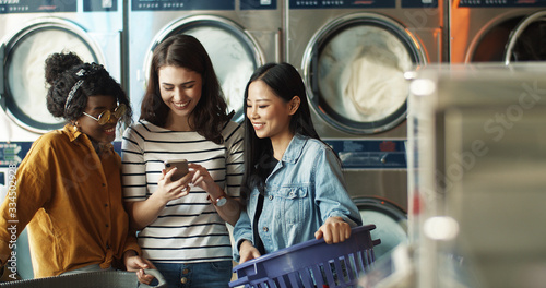 Slika na platnu Caucasian girl showing photos on smartphone to mixed-races female friends while washing machines working and cleaning clothes