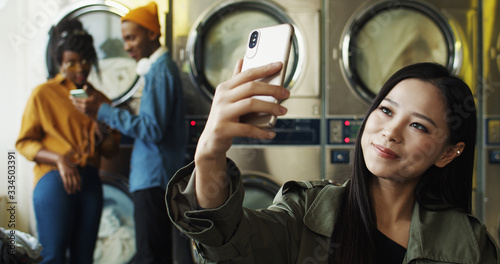 Asian young beautiful cheerful girl smiling and posing to smartphone camera while taking selfie photo in laundry service. Pretty happy woman making selfies photos with phone at washing machines.