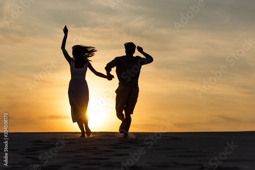 silhouettes of man and woman running on beach against sun during sunset