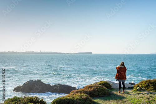 A person in the distance standing on the edge of a cliff with vegetation, looking at the ocean. View from the back.