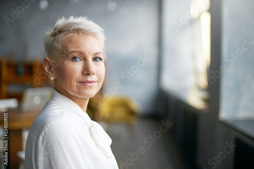 Isolated image of beautiful stylish middle aged female entrepreneur with neat make up going to business meeting, standing against office interior background, looking at camera with confident smile photo