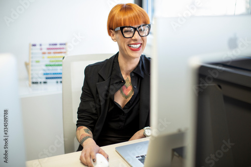 Ragazza tatuata con i capelli rossi e gli occhiali neri con un completo  nero ride nella sua postazione di lavoro photo