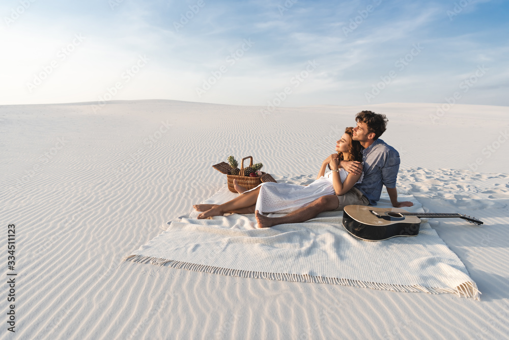 young couple hugging while sitting on blanket with basket of fruits and acoustic guitar on beach