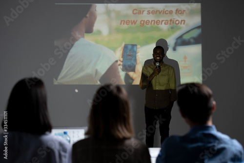 Cheerful Afro-American creative app developer standing against projection screen and presenting idea for car service to investors